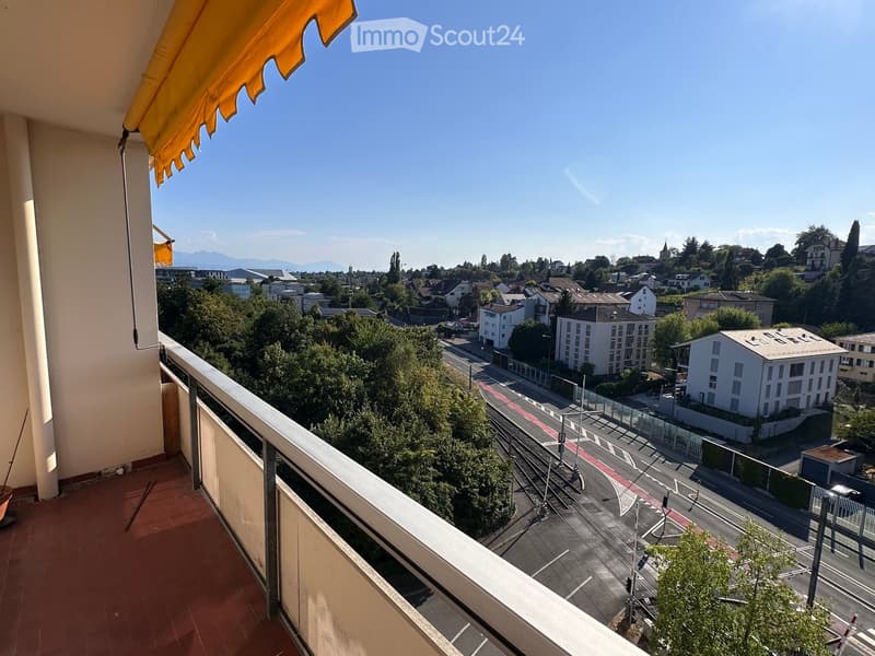 Balcony with a view of a cityscape, including buildings, trees, and a road below. Features a metal railing and an orange awning.