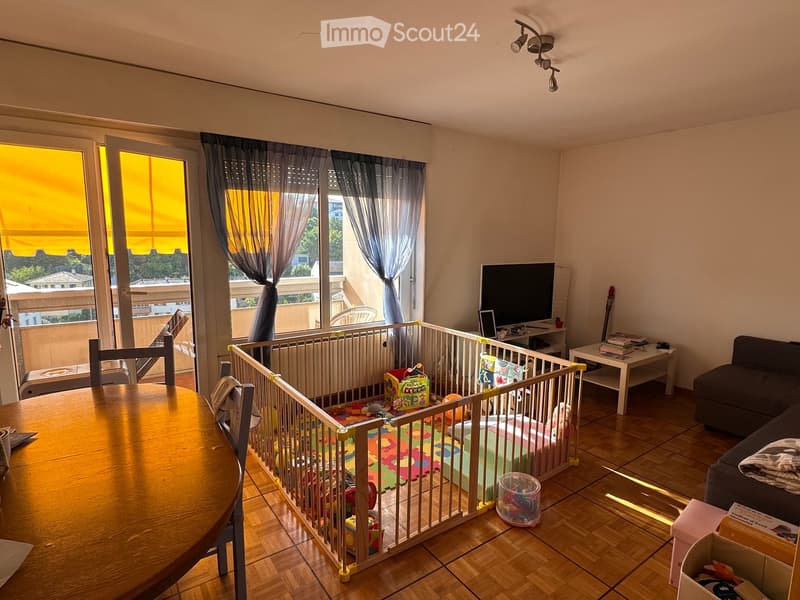 A well-lit living room with wooden flooring, a sofa, a dining table, a TV, and a playpen with toys.