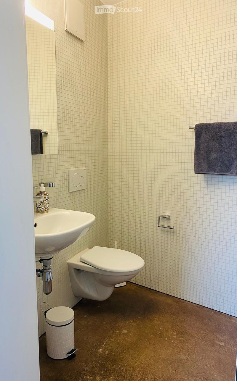 Modern bathroom with white tiled walls, a white sink, toilet, towel rack, and a small trash can on the floor.