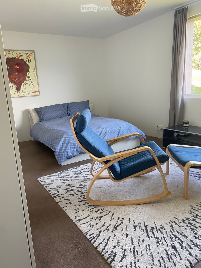Modern bedroom with a double bed, blue bedding, a wooden rocking chair, a small table, and a painting on the wall.