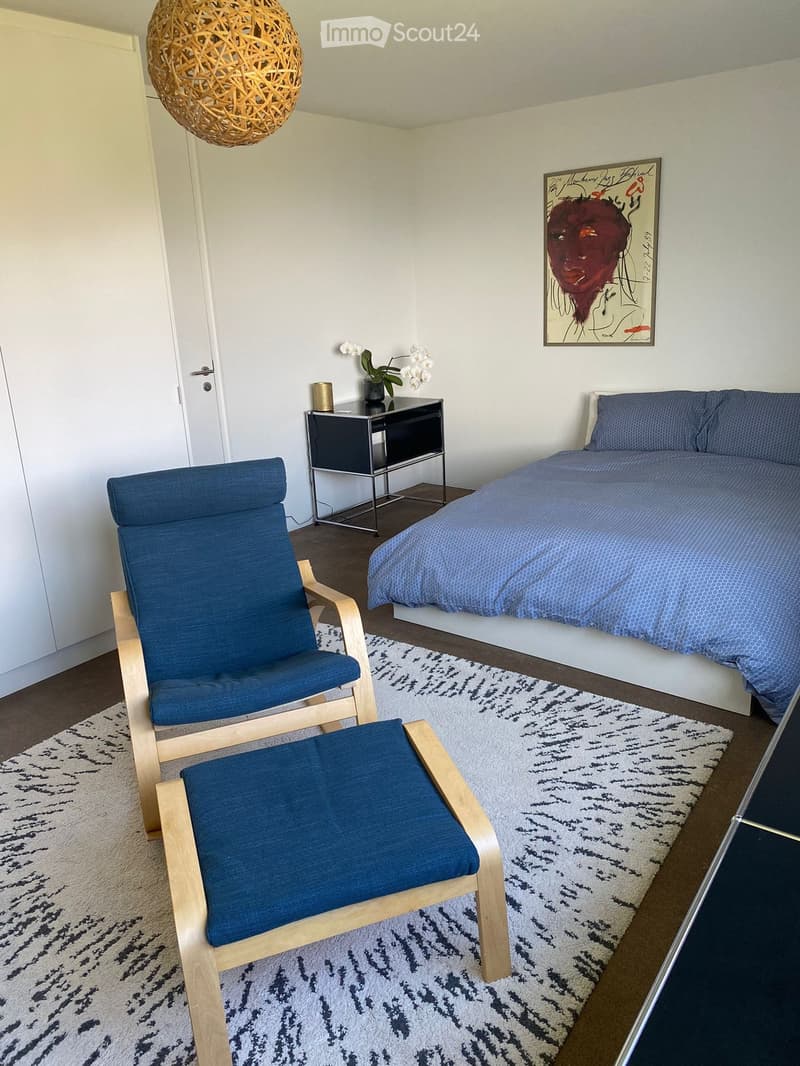 Modern bedroom with a blue duvet-covered bed, wooden chair and stool, nightstand with vase and lamp, and a colorful abstract painting on the wall.