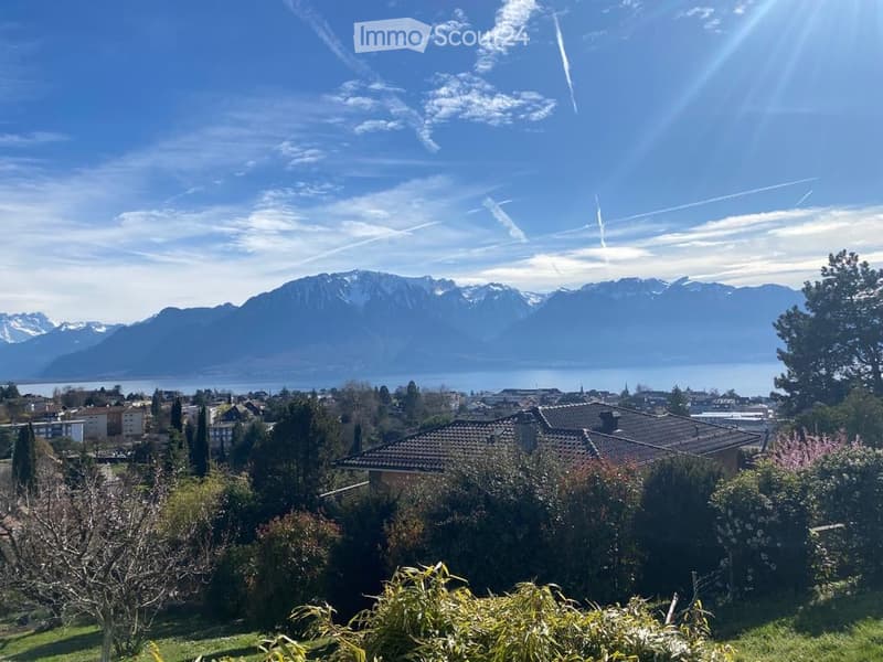 Panoramic view of a cityscape with mountains and a lake in the background, surrounded by trees and greenery.