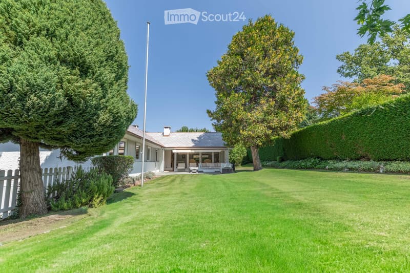A well-maintained garden with a lawn, trees, and a modern house with a patio and outdoor seating area.