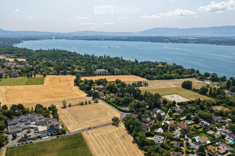Aerial view of a suburban area with houses, fields, and a large body of water in the background. The area includes residential buildings, agricultural fields, and a waterfront with boats.