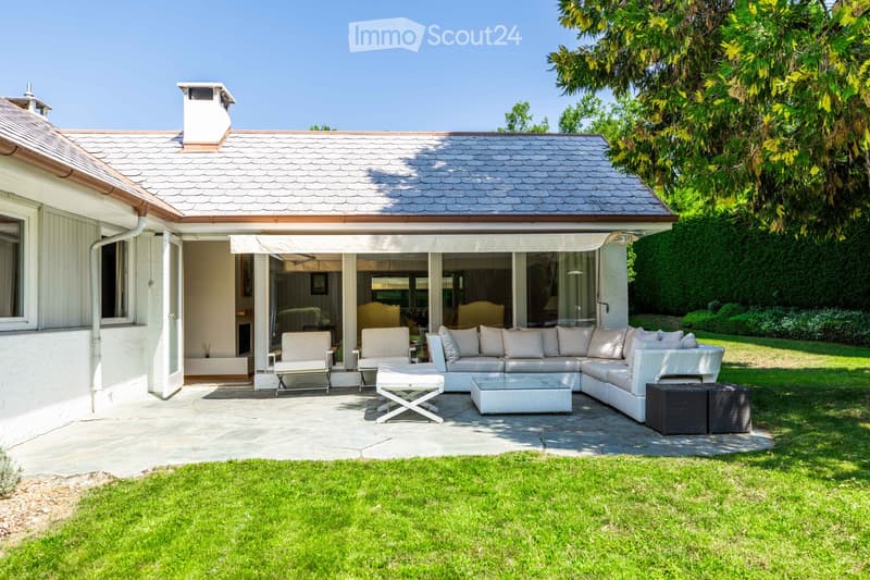 A modern outdoor patio area with a large white sectional sofa, two white chairs, a small table, and two black stools, surrounded by a well-maintained lawn and lush greenery.