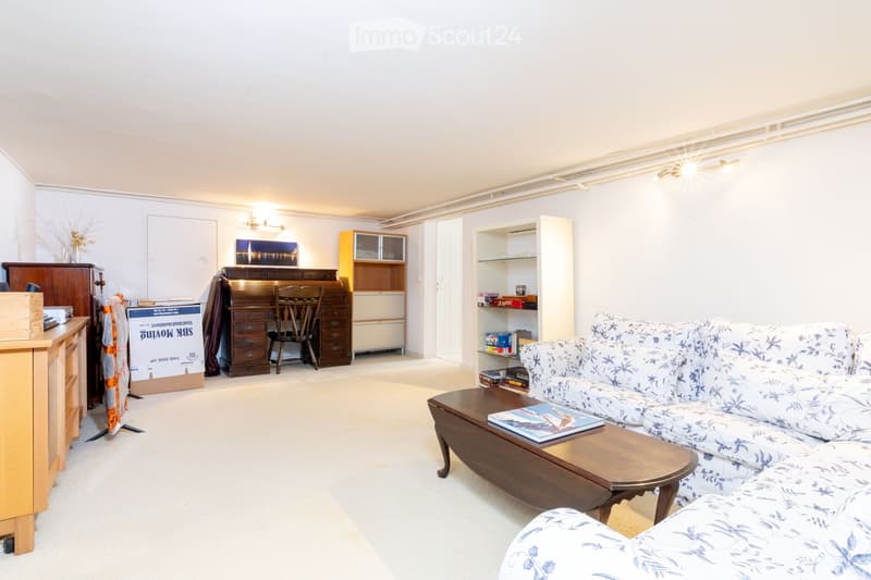 A well-lit living room with a patterned sofa, coffee table, wooden cabinet, bookshelf, and a desk area with a chair.