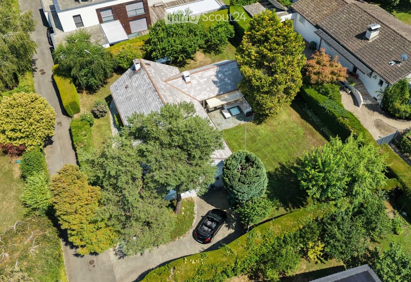 Aerial view of a residential house with a well-maintained garden, driveway, and surrounding greenery. The house features a tiled roof and is surrounded by trees and shrubs.
