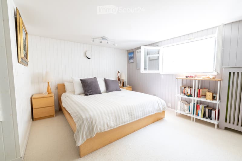 Cozy bedroom with a wooden bed, nightstand, bookshelf, and decorative items. The room features white shiplap walls and a large window with blinds.
