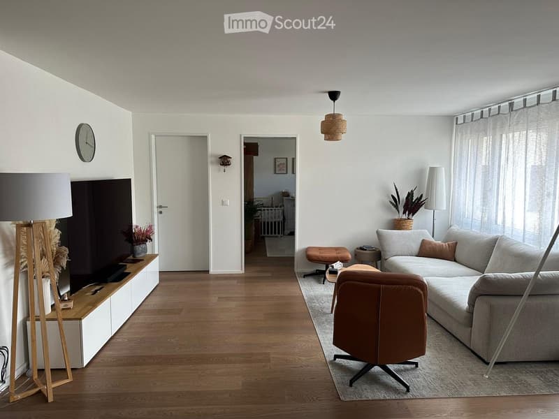 Modern living room with light-colored walls, wooden flooring, and large windows. Features a grey sofa, brown leather chair, coffee table, floor lamp, and decorative plants.