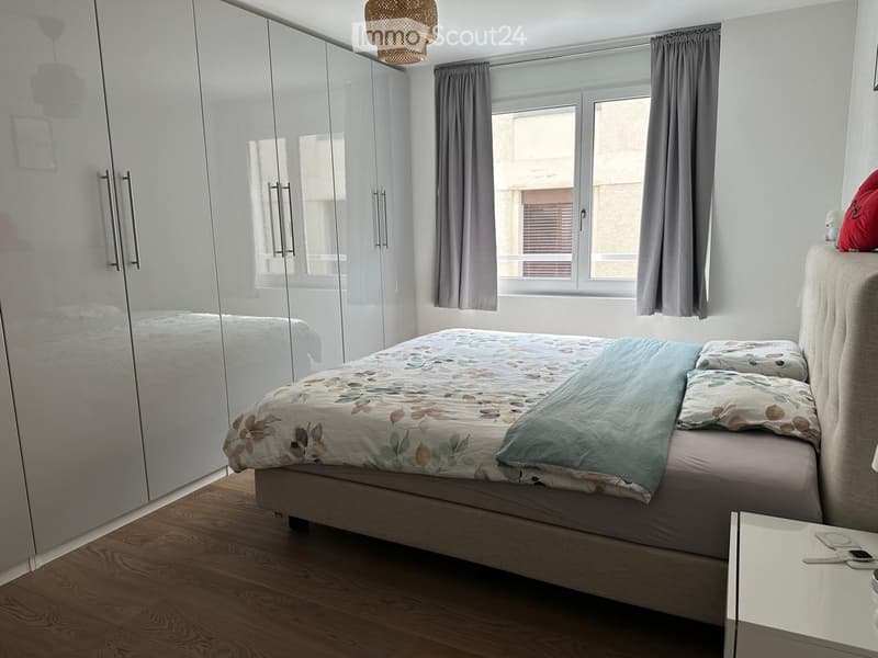 Modern bedroom with a large bed, floral duvet, nightstand, and mirrored wardrobe. Large windows with grey curtains provide natural light.