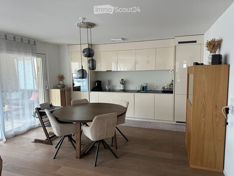 Modern kitchen and dining area with light-colored cabinets, a round wooden dining table with four chairs, and various kitchen appliances on the counter.