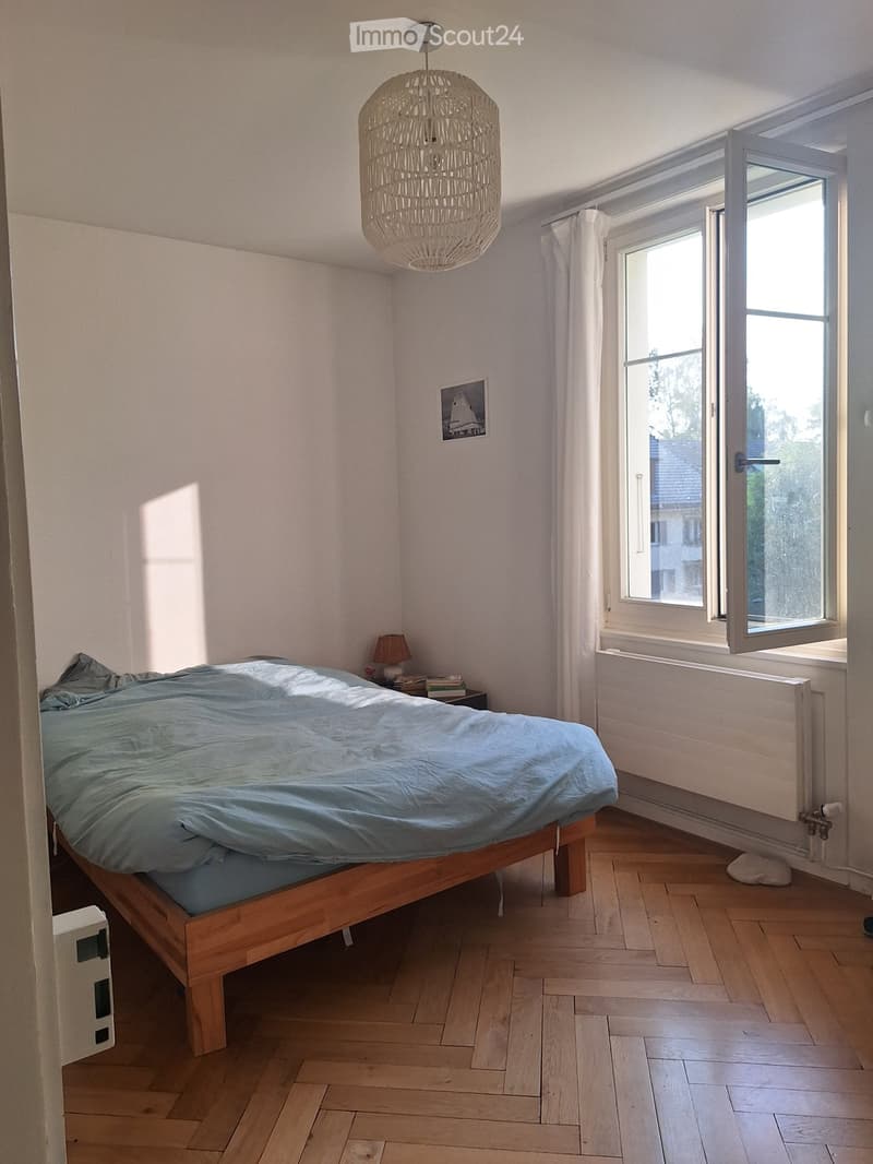 A well-lit bedroom with wooden flooring, a large bed with blue bedding, a nightstand with a lamp, and a picture frame on the wall. A large window with white curtains allows natural light to enter.
