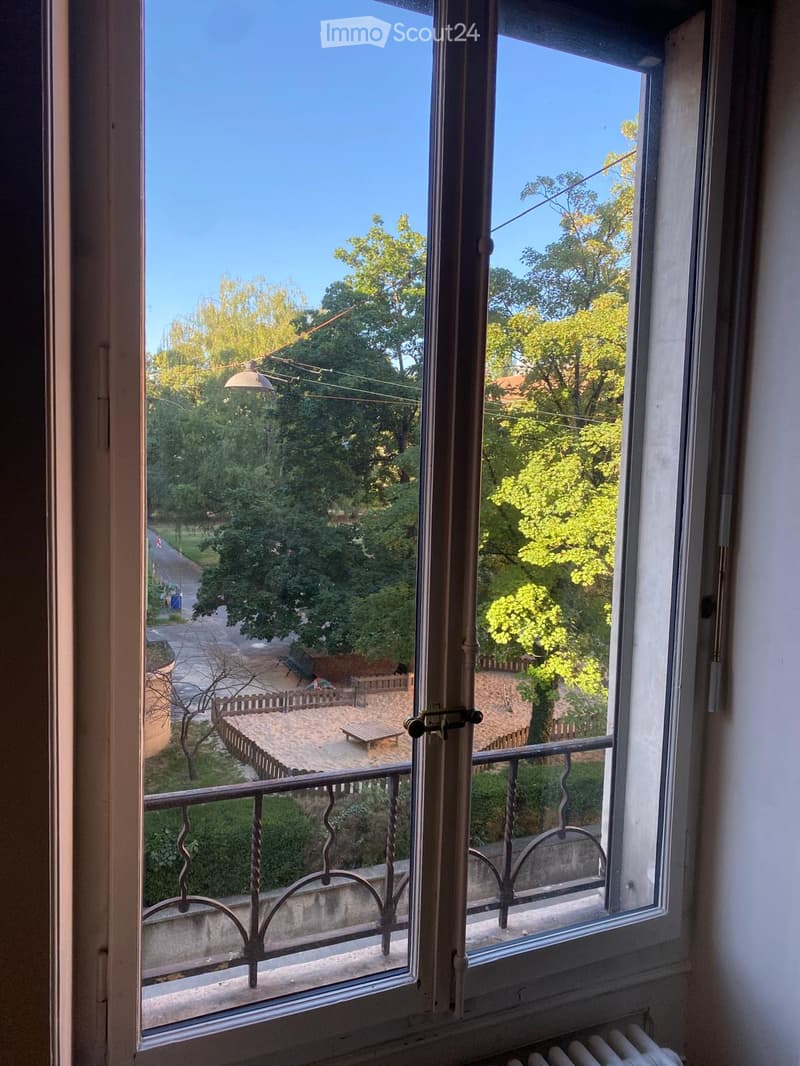 Balcony with metal railing overlooking a green area with trees, a bench, and a pathway.