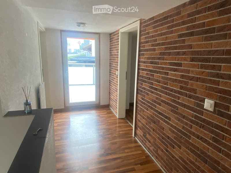 Hallway with wooden flooring, brick wall on one side, and a door leading outside. A small shelf with decorative items is visible on the left.