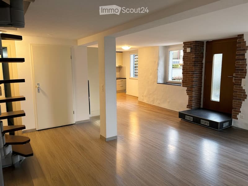 Empty room with wooden flooring, white walls, and a staircase leading to another level. The room has a large window with a view of the outside and a partially visible kitchen area in the background.