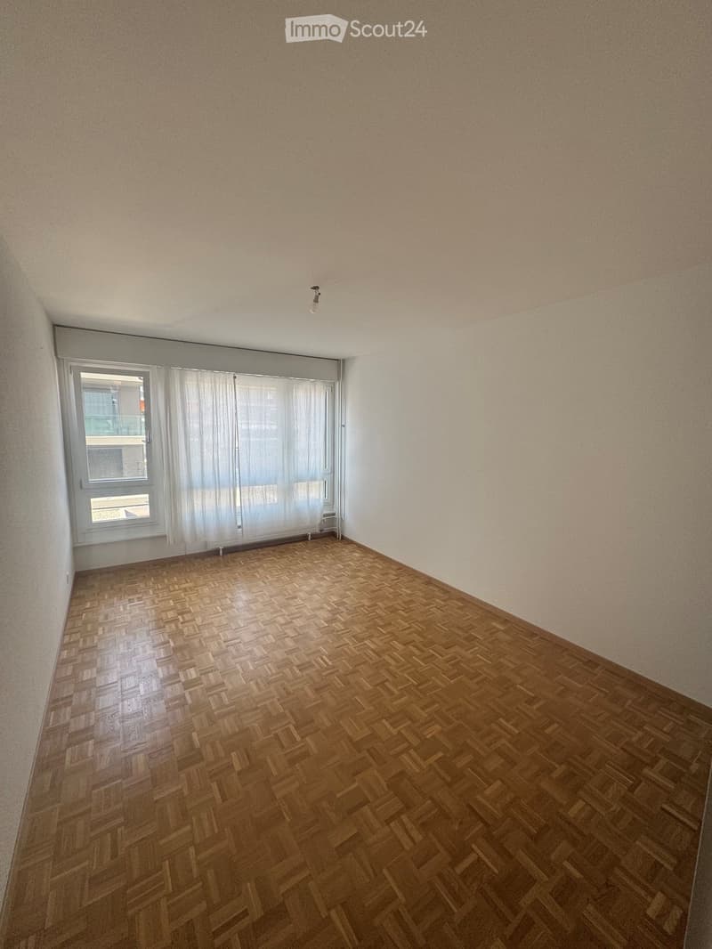 Empty room with wooden herringbone flooring, white walls, and a large window with white curtains.