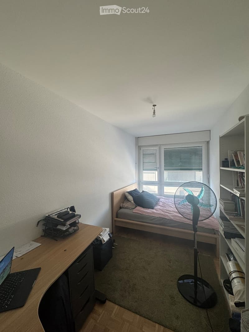 A small, furnished bedroom with a bed, desk, laptop, fan, and bookshelf.