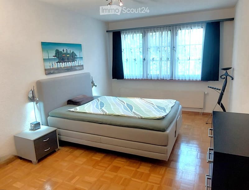 Modern bedroom with a large bed, nightstand, lamp, and a painting on the wall. The room features wooden flooring and large windows with curtains.