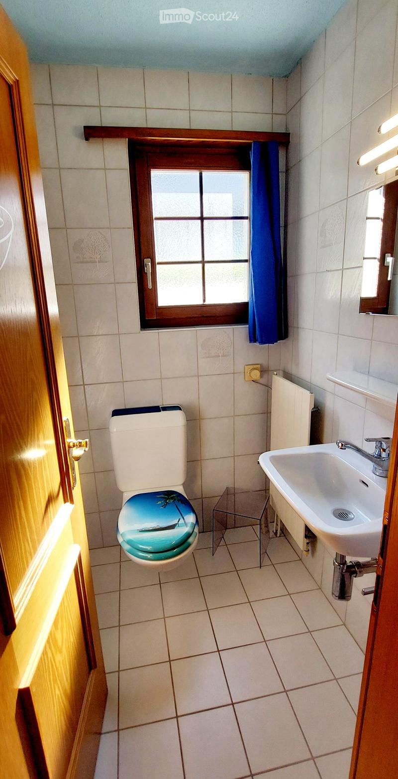 Bathroom with white tiled walls and floor, a toilet with a decorative seat cover, a sink with a faucet, and a small glass stool.