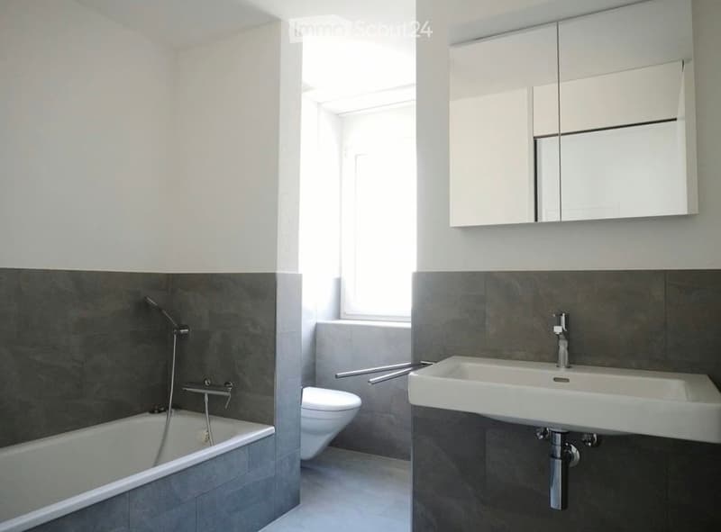 Modern bathroom with a bathtub, toilet, sink, mirror, and cabinet. The walls are painted white with a dark grey lower half, and the floor is tiled in light grey.