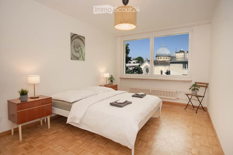 Bedroom with a bed, nightstands, lamps, a chair, a radiator, a window with a view of a dome building, and a picture frame on the wall.