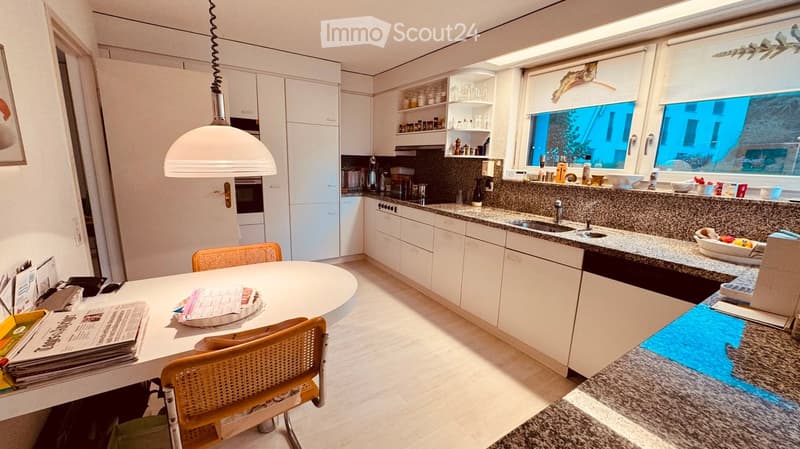 Modern kitchen with white cabinets, granite countertops, a round dining table with two chairs, and various appliances including an oven, dishwasher, and sink. The space is well-lit with natural light from large windows and a pendant light hanging above th