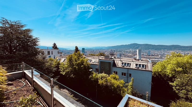 A rooftop terrace with a glass railing, overlooking a cityscape with buildings and trees under a clear blue sky.