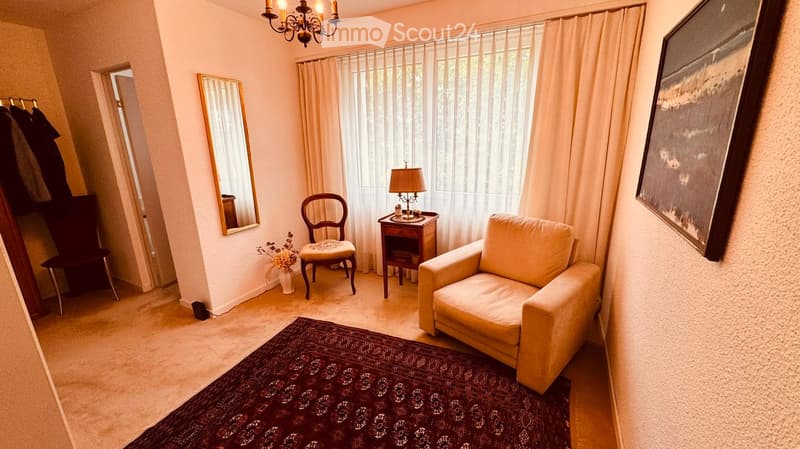 A cozy living room with a beige sofa, wooden chair, small wooden table with a lamp, a large mirror, and a patterned rug on the floor.