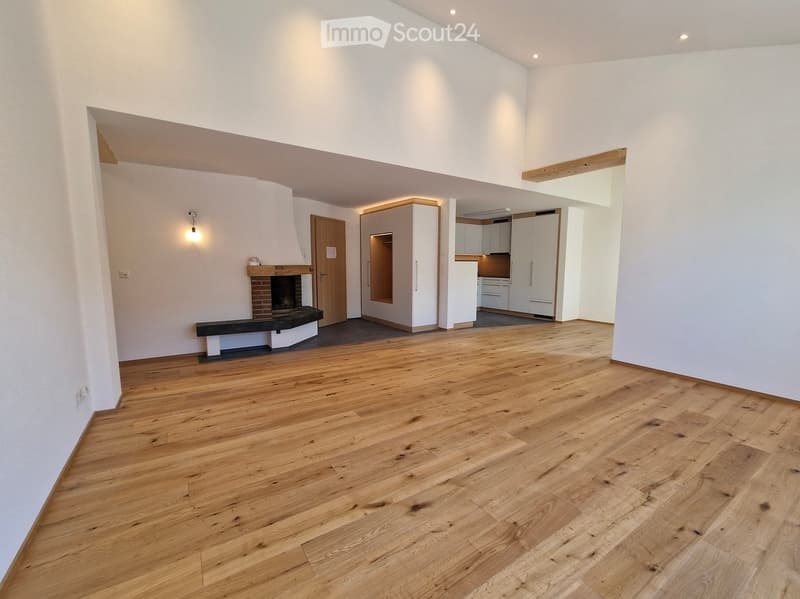Open-plan living space with wooden flooring, white walls, fireplace, and adjacent kitchen area with white cabinets and appliances.