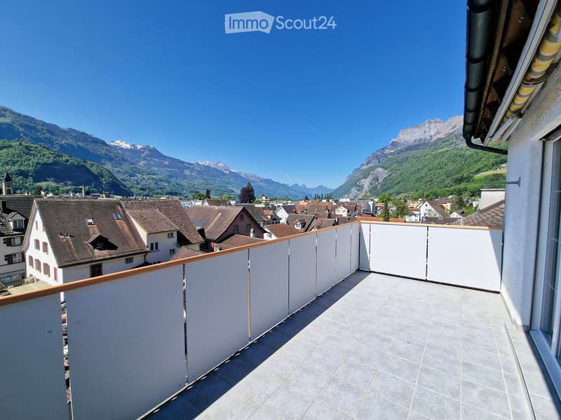Balcony with tiled floor, white walls, wooden railing, overlooking a town with houses and distant mountains.