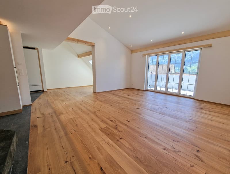 Empty room with wooden flooring, white walls, and large windows with a view of greenery outside.