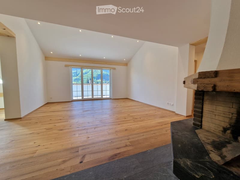 Empty room with wooden flooring, white walls, and a fireplace. Large window with a scenic view.