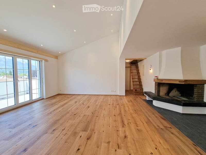 Empty living room with wooden flooring, white walls, large windows, and a fireplace with a wooden mantle.