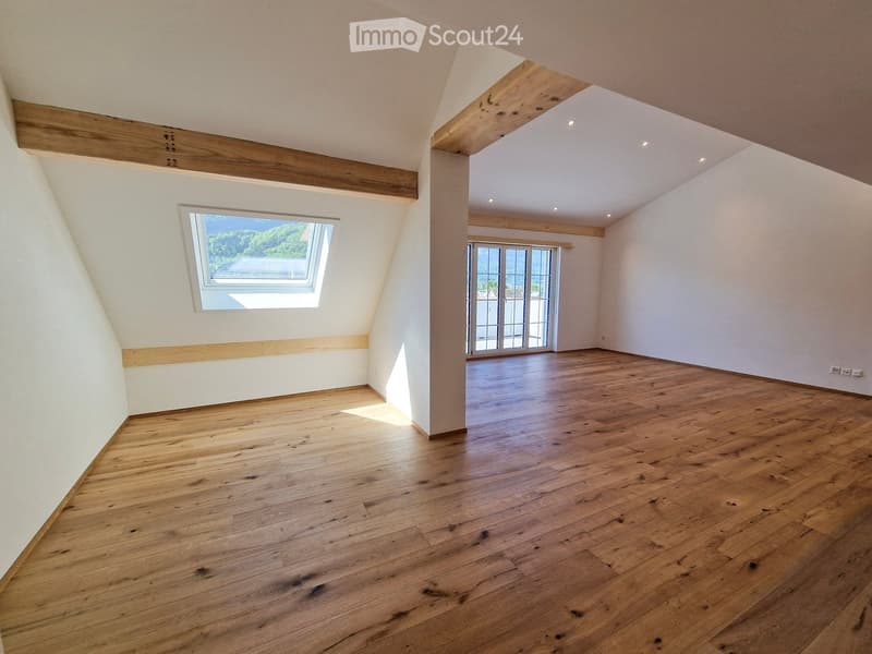 Empty attic room with wooden floors, white walls, exposed wooden beams, skylight window, and sliding glass door leading to a balcony.