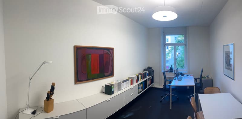 Modern office space with white walls, large window, desk with computer, bookshelf, and artwork.