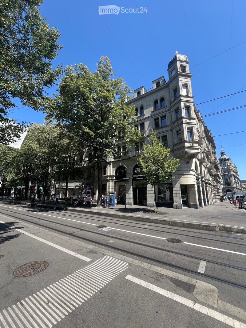 A multi-story building with classical architecture, featuring arched windows, a domed roof, and ornate detailing. The building is situated on a street with a sidewalk, trees, and a pedestrian crossing.