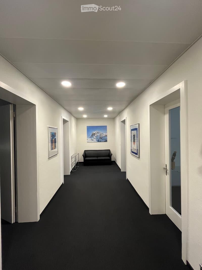 A narrow hallway with white walls, black carpet, and a small black sofa placed against the wall. There are picture frames on the walls and a glass door at the end.