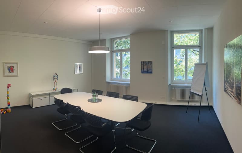 Bright conference room with a round table surrounded by black chairs, a large screen on a stand, framed artwork, and a colorful sculpture on the floor.
