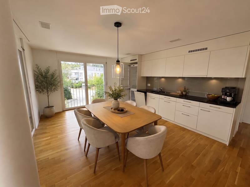 Open-plan kitchen and dining area with light wood flooring, white cabinets, a central dining table with six chairs, and a coffee machine on the counter.