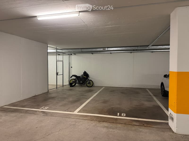 Underground parking area with concrete floors, white walls, and numbered parking spaces. A motorcycle is parked in one space, and a car is partially visible in another. The area is well-lit with fluorescent lighting.