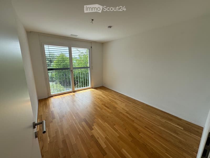 Empty room with light wooden flooring, white walls, and a large sliding glass door leading to a balcony with a metal railing.