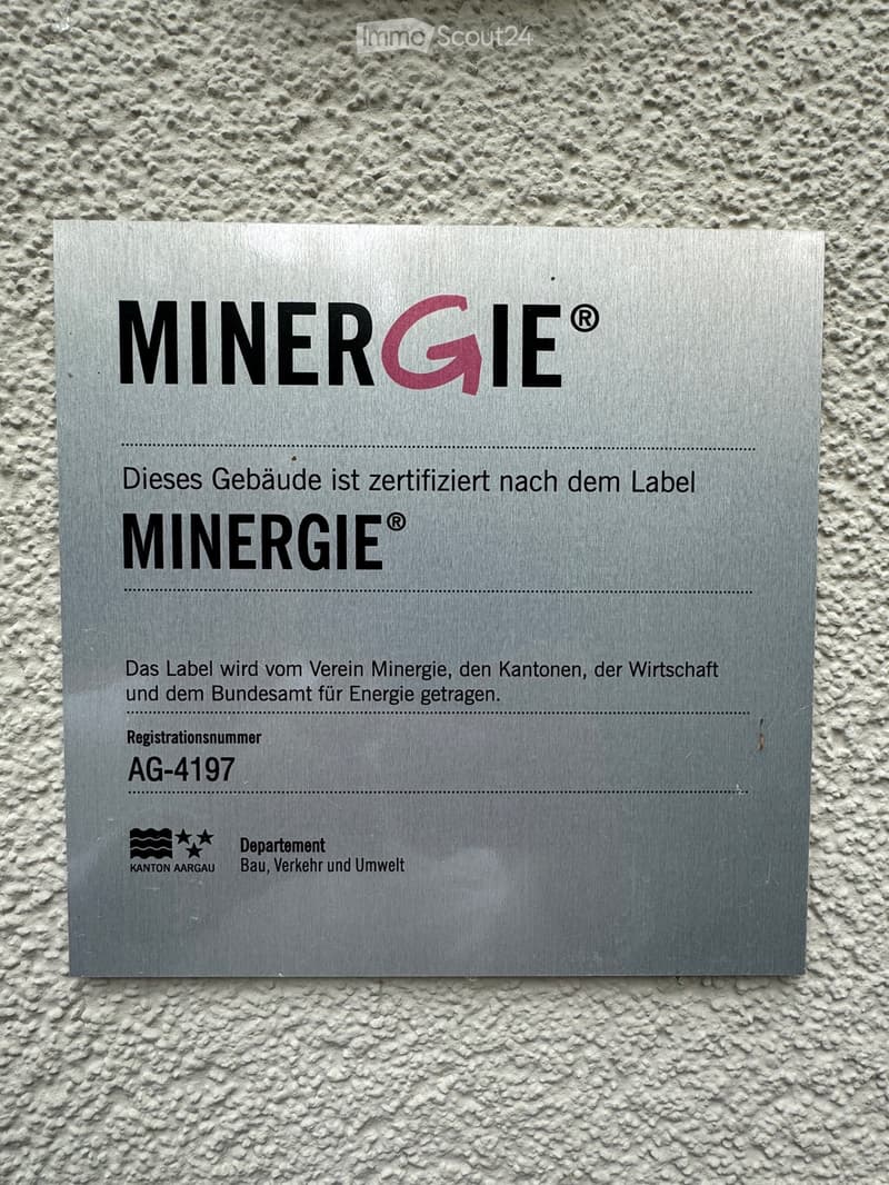 A silver plaque mounted on a textured wall, displaying the Minergie certification logo and details about the building's energy certification.