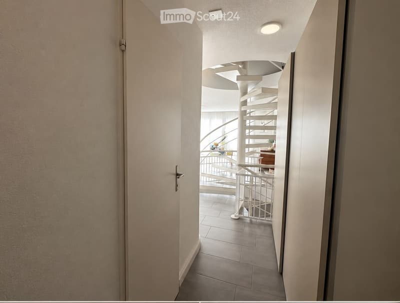 A modern hallway with a curved staircase visible in the background, featuring white walls, tiled flooring, and recessed lighting.