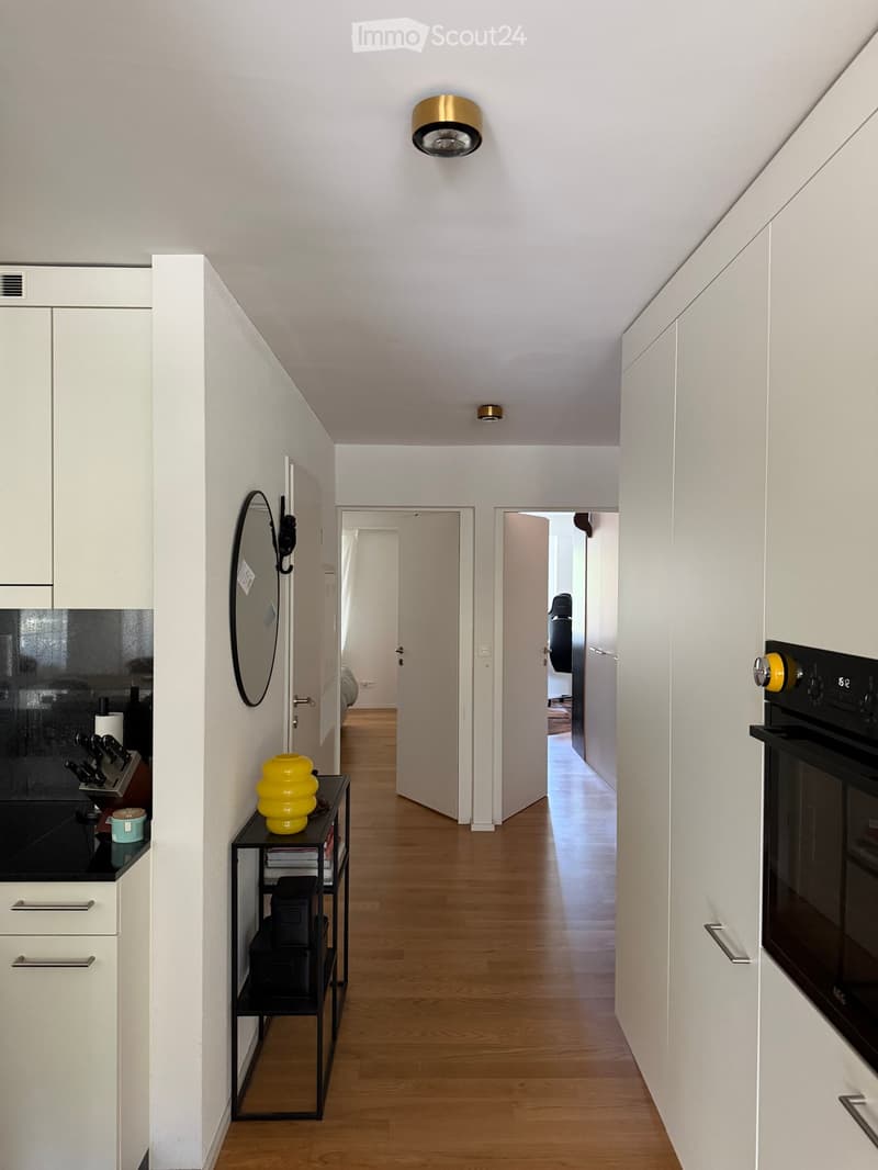 Modern hallway with light wooden flooring, white walls, and built-in white cabinets. Features a small black side table with a yellow vase, a mirror on the wall, and an oven on the right side.