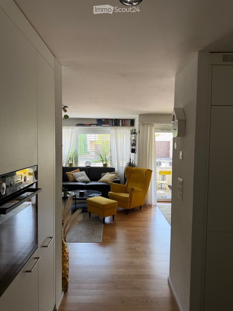 Modern living room with hardwood floors, large windows, black leather sofa, yellow armchair, ottoman, coffee table, and decorative vase.