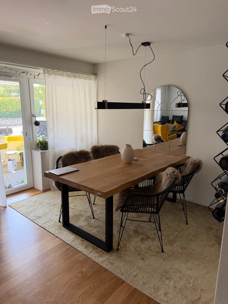 Modern dining area with a wooden table, black metal chairs with fur cushions, a laptop, a vase, and a large mirror. The space features a pendant light fixture and a view of a garden through large windows.