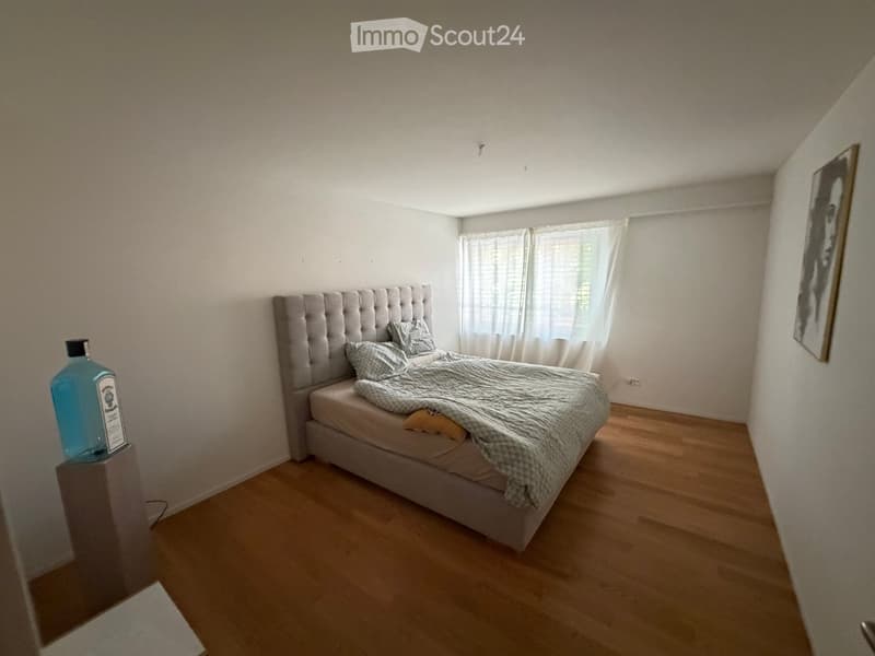 A modern bedroom with a tufted headboard bed, pillows, a blanket, a blue bottle on a nightstand, and a framed picture on the wall. The room features wooden flooring and large windows with white curtains.