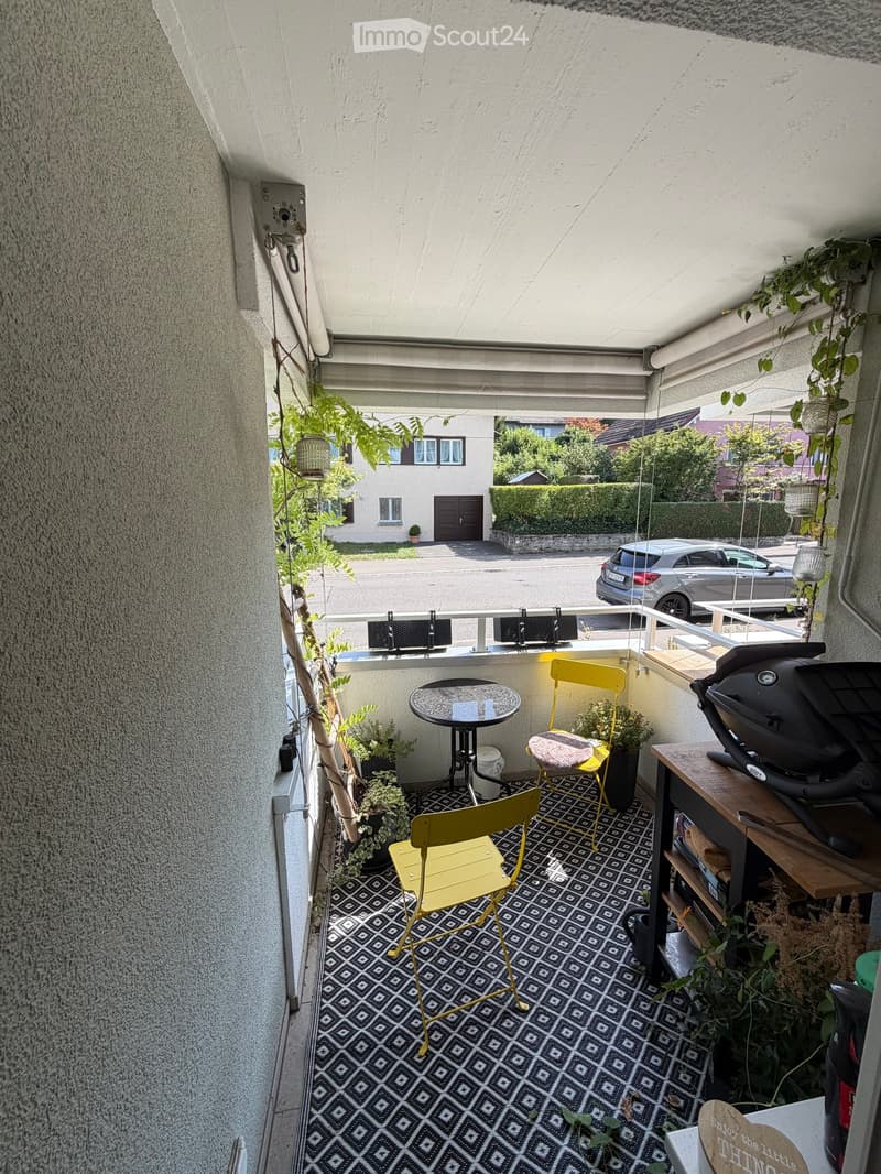 Balcony with a small round table, two yellow chairs, a grill, and several potted plants. The floor has a patterned tile design.