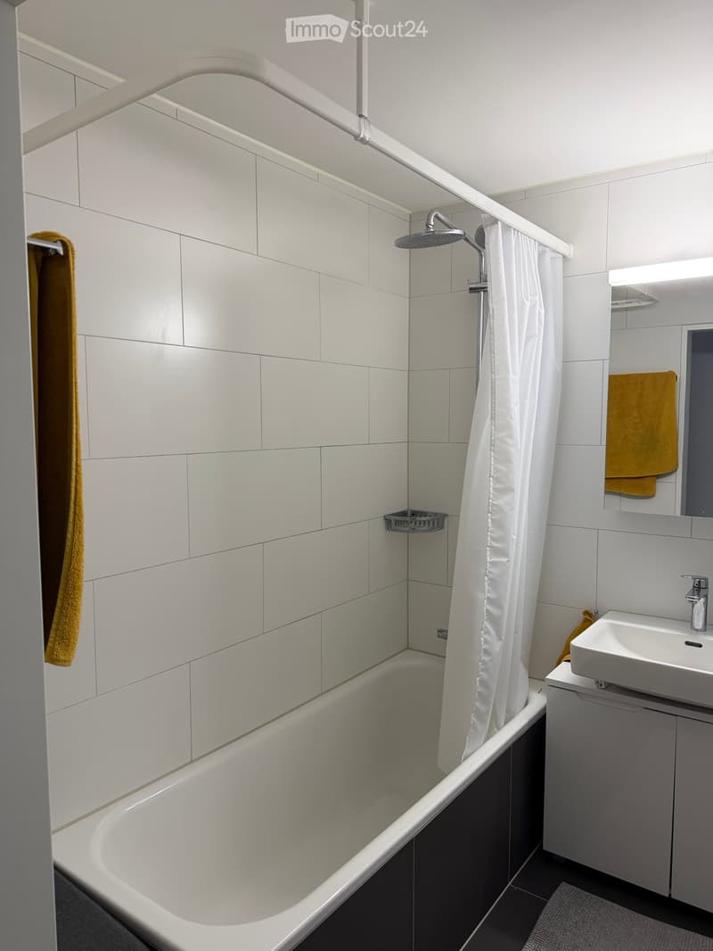 Modern bathroom with a white bathtub, shower, sink, and mirror. White tiles and a white shower curtain are visible.