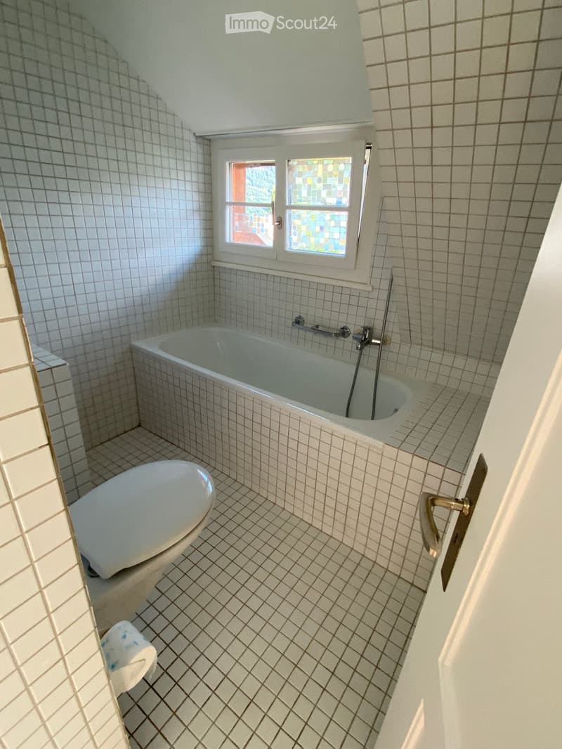 Bathroom with white tiled walls and floor, featuring a bathtub, toilet, and a window with colorful stained glass. The door handle is visible on the right side.
